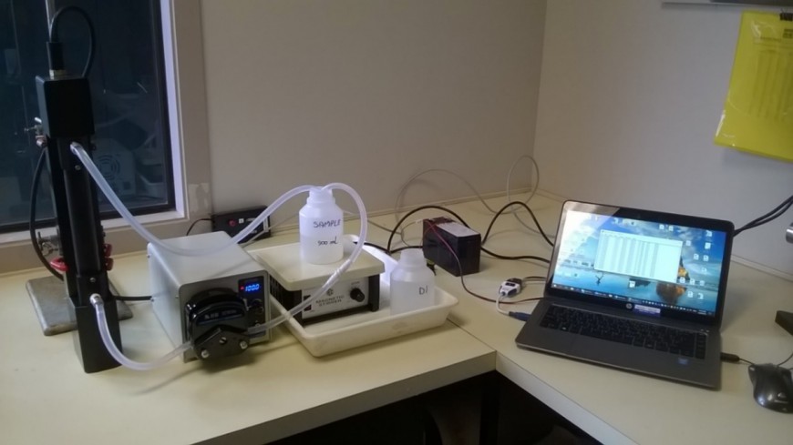 Figure 2: WET Labs® C-Star beam transmissometer set up in flow-through mode for laboratory measurements of beam attenuation coefficient (beam-c). Left to right: C-Star sensor (black item) with flow-tube fitted, peristaltic pump, diluted sample (stirred by magnetic stirrer), and laptop computer running Terra Term web 3.1. The C-Star sensor emits a collimated beam of green light (530 nm) and measures the fraction transmitted through the water sample.